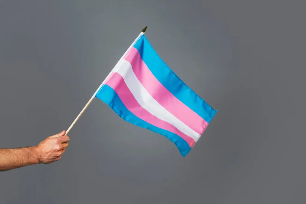Photo of a hand holding a trans pride flag (Blue, pink, white, pink, blue). Hormone replacement.